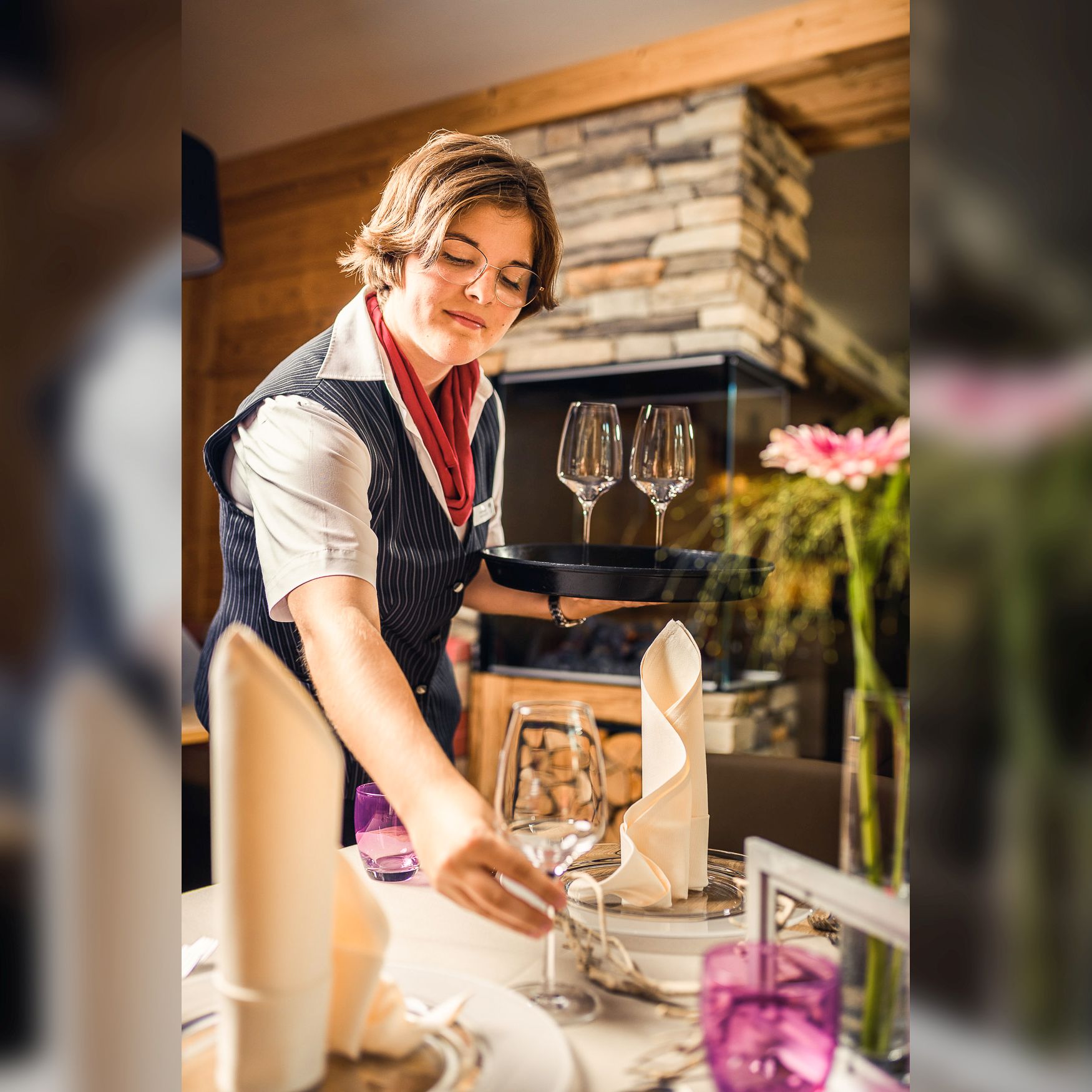 Eine Servicemitarbeiterin mit kurzen Haaren und Brille, gekleidet in eine gestreifte Weste und ein rotes Halstuch, deckt einen Tisch im Restaurant "Seinerzeit" ein. Sie hält ein Tablett mit Weingläsern in der linken Hand und platziert ein Glas auf dem Tisch mit der rechten Hand. Der Tisch ist elegant gedeckt mit weißen Stoffservietten, lila Gläsern und einem Blumenarrangement im Hintergrund. Ein Kamin mit Steinverkleidung trägt zur gemütlichen Atmosphäre bei.