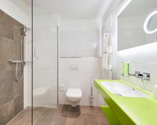 Helles Badezimmer im Parkhotel Neustadt mit weißen Fliesen und einer lebhaften, grünen Ablagefläche. Zur Ausstattung gehören ein weißes Waschbecken, ein großer Spiegel mit integrierter Beleuchtung, eine ebenerdige begehbare Dusche mit modernen Armaturen und eine Toilette. Frische Handtücher sind an einem Handtuchhalter ordentlich platziert.